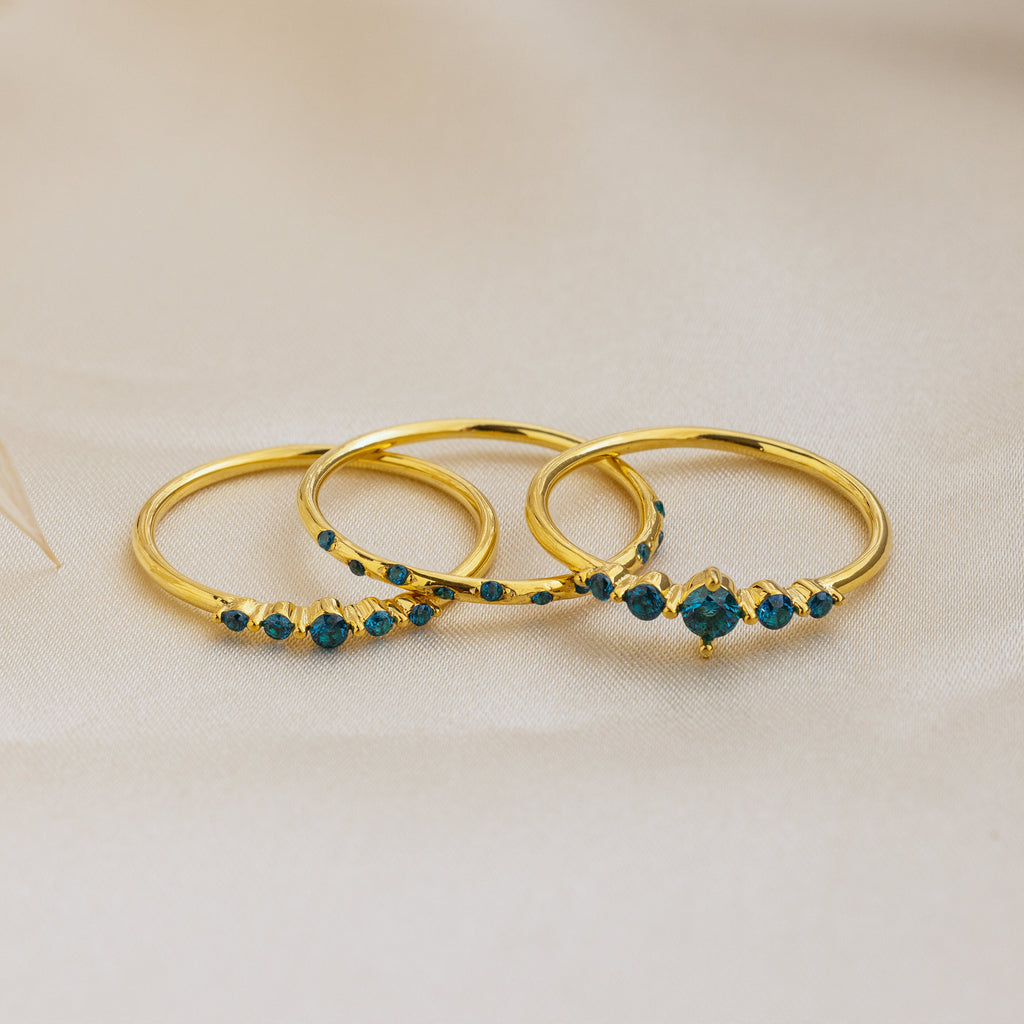 The Lace Birthstone Rings Set features three delicate rings with small blue gemstones, displayed on soft beige fabric—a perfect personalized gift for lovers of minimalist jewelry.