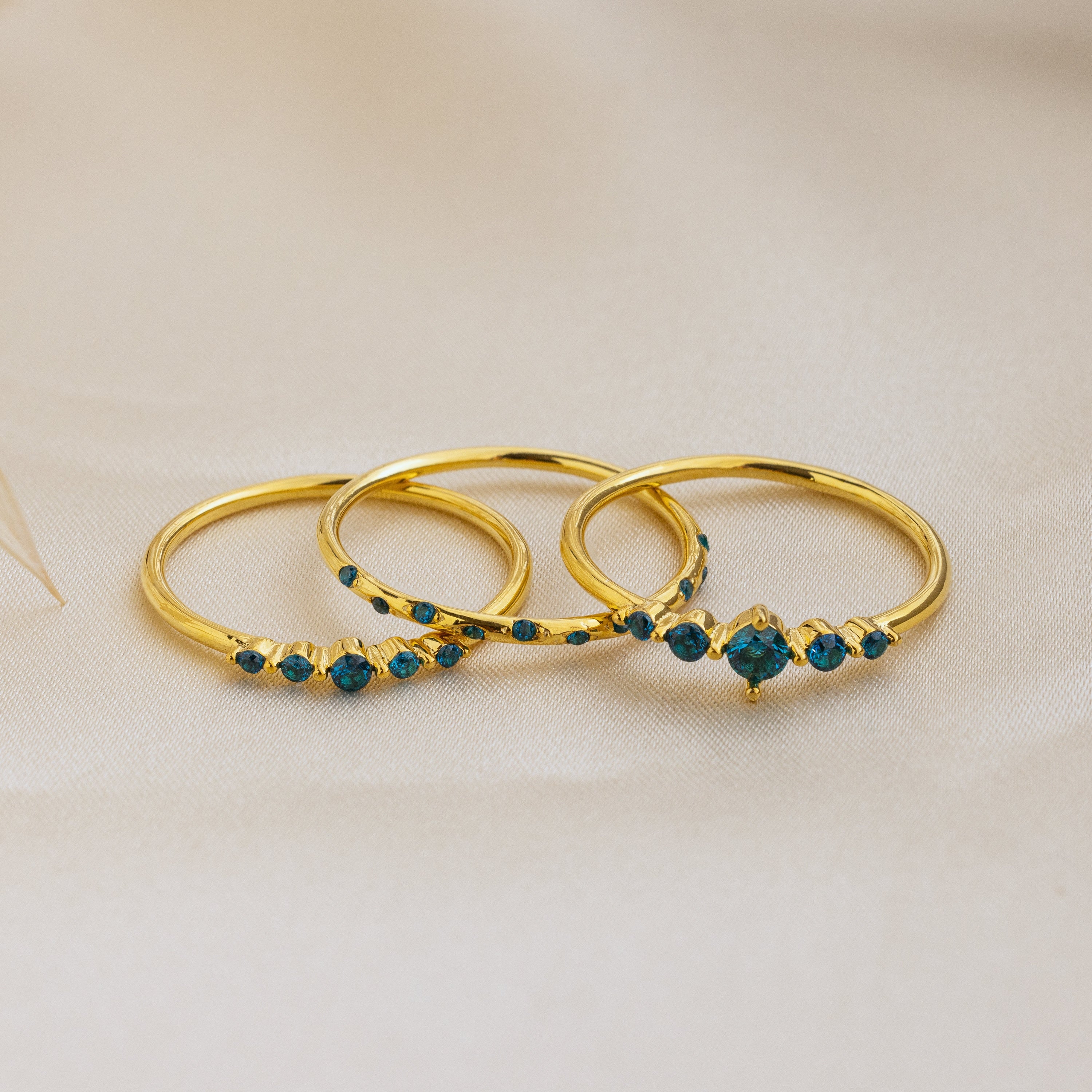 The Lace Birthstone Rings Set features three delicate rings with small blue gemstones, displayed on soft beige fabric—a perfect personalized gift for lovers of minimalist jewelry.