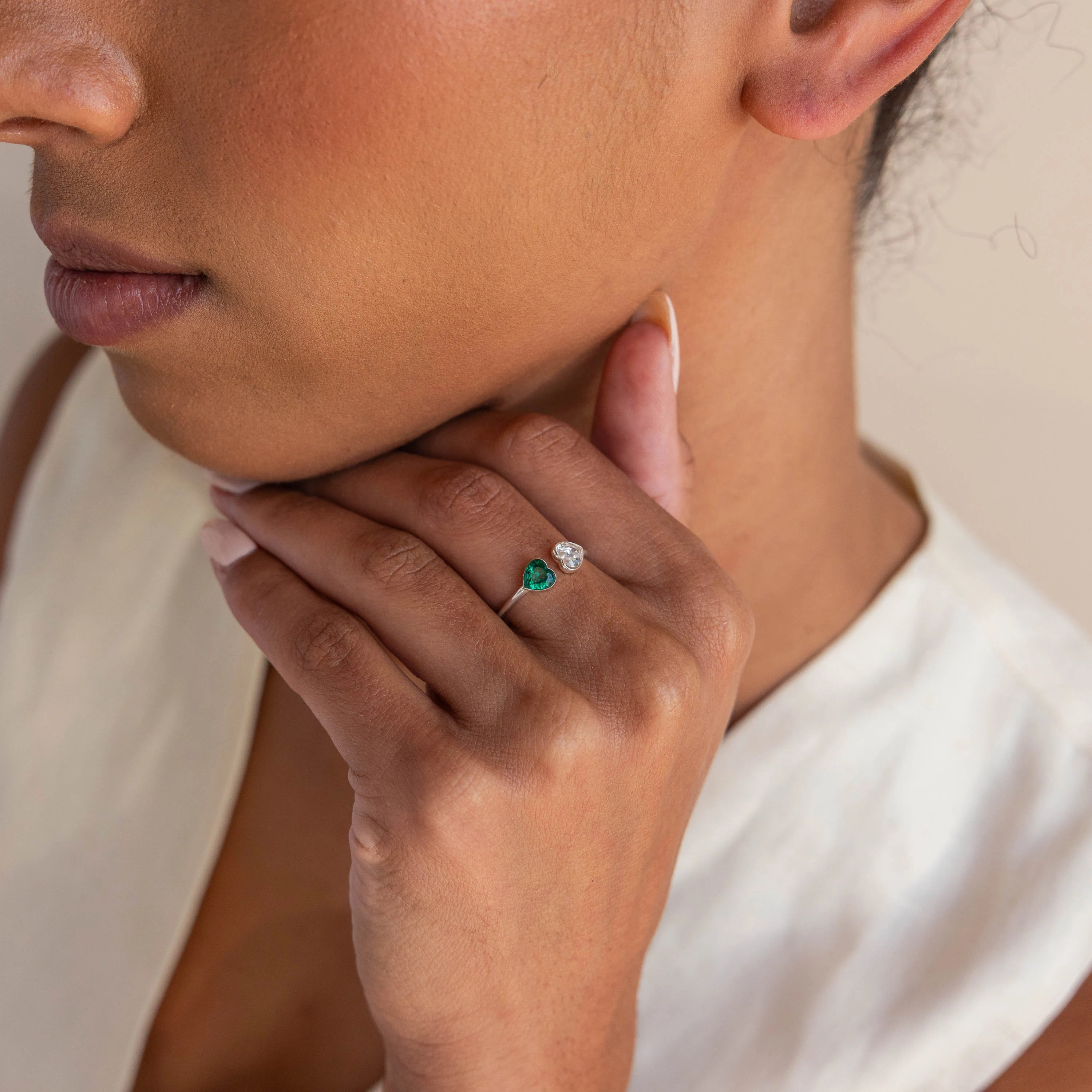 A woman touches her neck while wearing the Duo Heart Birthstone Ring, featuring green and clear heart-shaped gemstones on her finger.