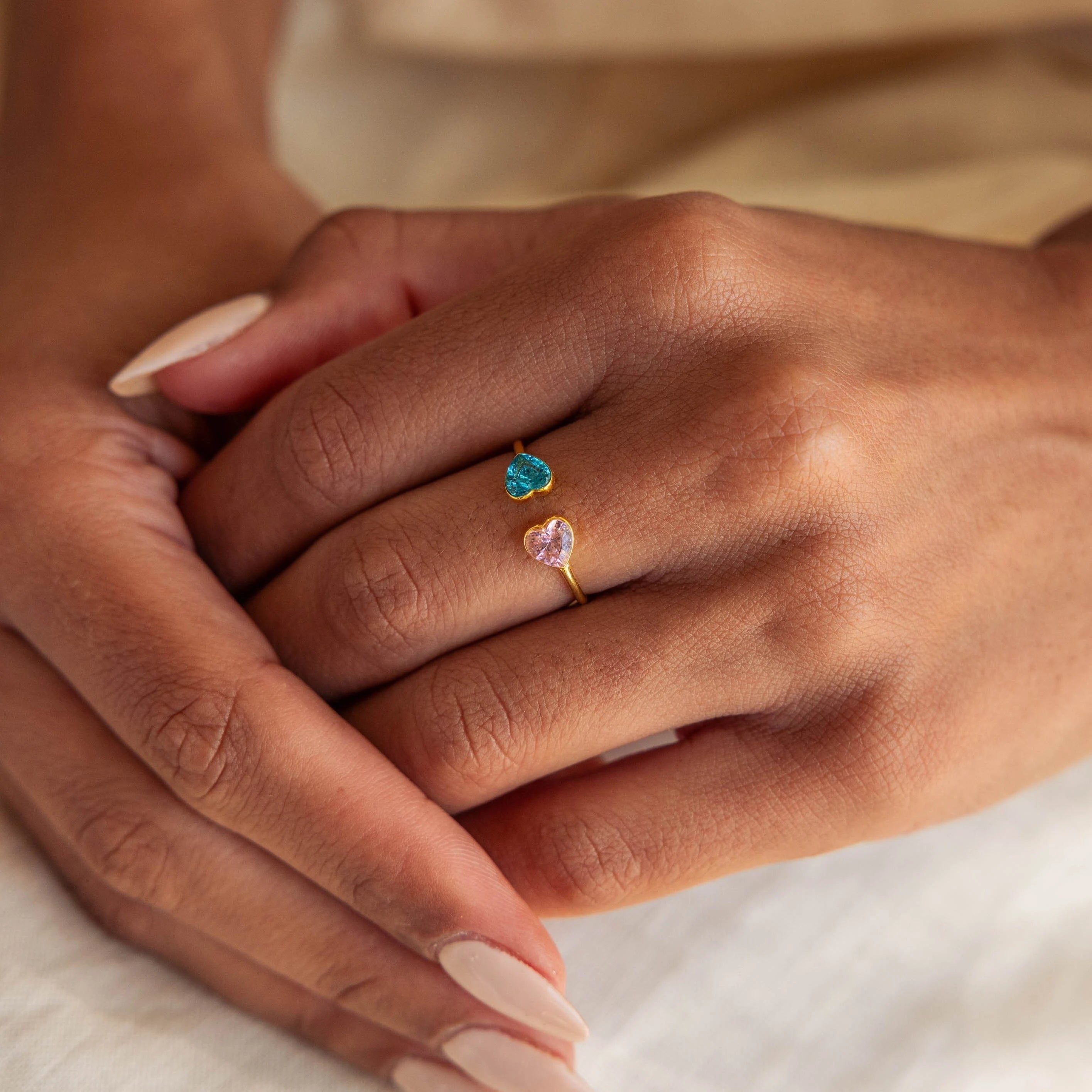 A hand with light beige nails wears the Duo Heart Birthstone Ring, featuring gold and blue and pink heart-shaped gemstones.