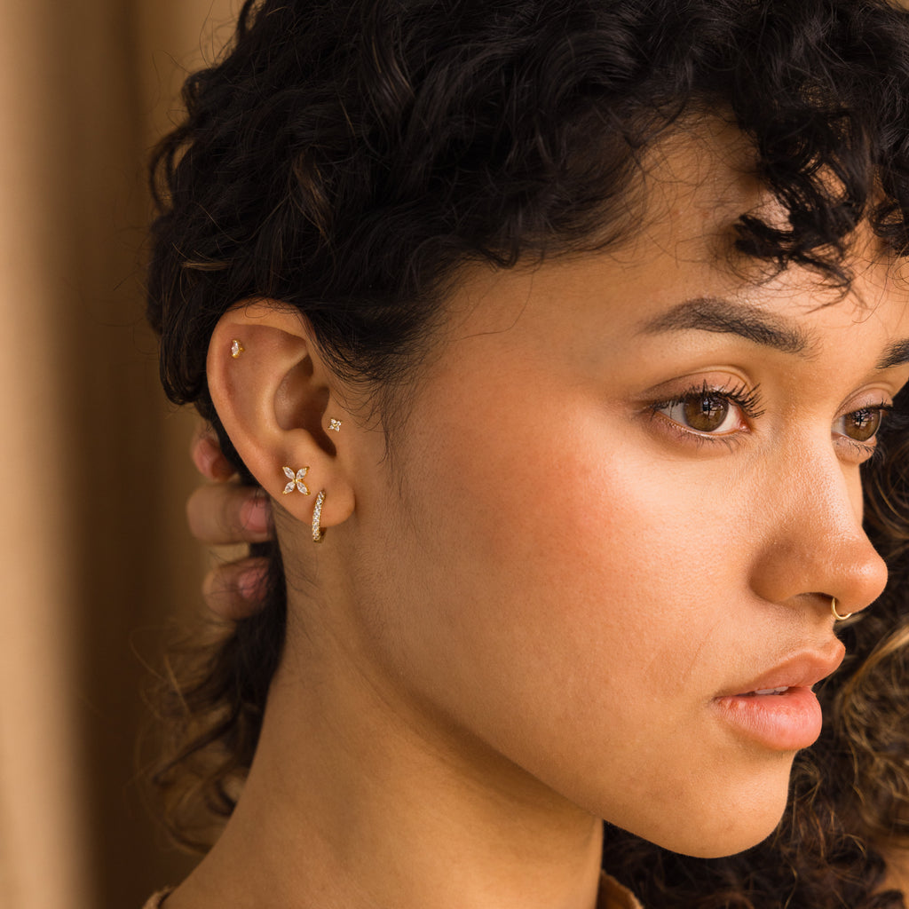 A woman with curly hair wearing multiple gold earrings, including Marquise Birthstone Studs, and a gold nose ring, looking to the side.