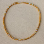 The Opal Bezel Tennis Necklace features small, round opals set in a single row, displayed on a beige textured surface.