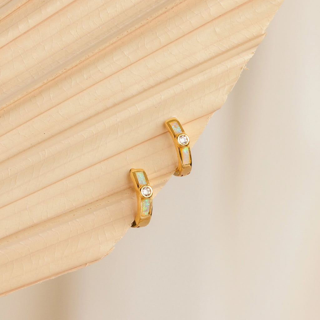 Opal Inlay Diamond Huggies shimmer with gemstone accents as they rest gracefully on a beige, textured palm leaf background.