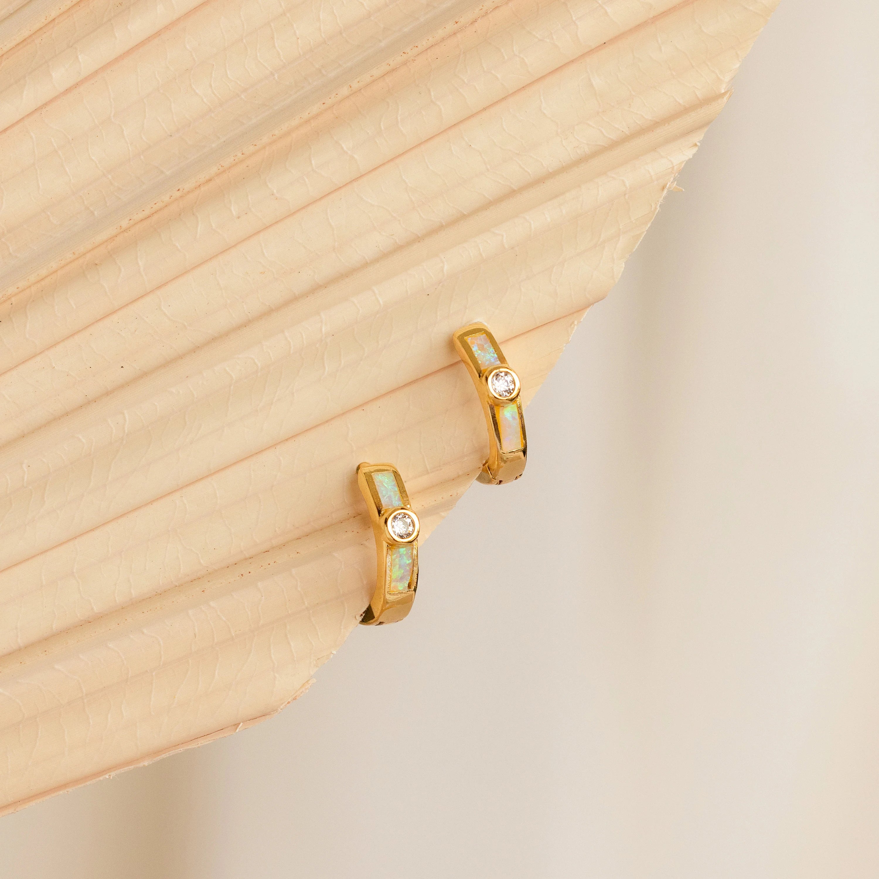 Opal Inlay Diamond Huggies shimmer with gemstone accents as they rest gracefully on a beige, textured palm leaf background.