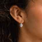 Close-up of a woman's ear wearing Round Pearl Drop Earrings, featuring a gold design with a white pearl-like oval pendant for an elegant, timeless look.