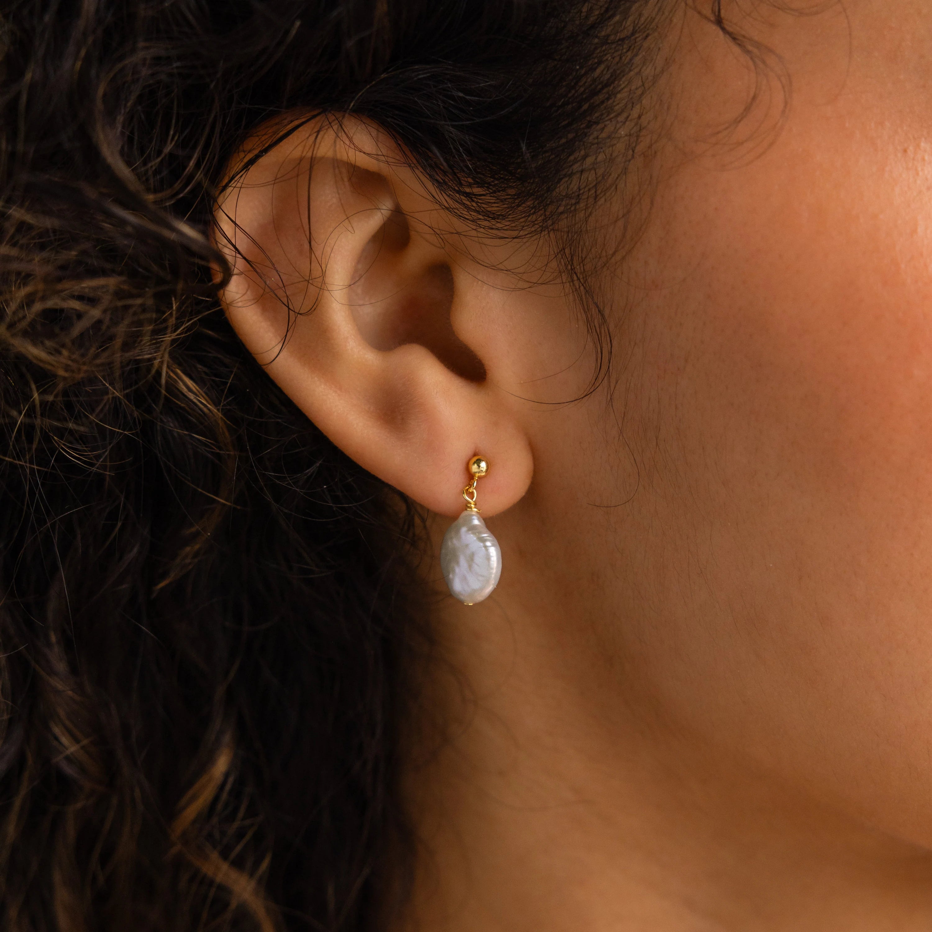 Close-up of a woman's ear wearing Round Pearl Drop Earrings, featuring a gold design with a white pearl-like oval pendant for an elegant, timeless look.