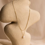 The Duo Chain Pearl Necklace features elegant pearls and a single pendant, showcased on a beige abstract bust with a soft neutral background. This timeless piece is perfect for wearing alone or layered with other necklaces.
