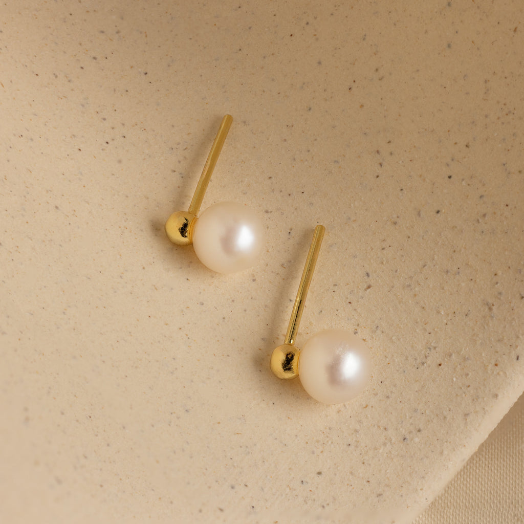 Classic Pearl Studs displayed on a textured beige surface, perfect for those who appreciate dainty earrings with timeless elegance.