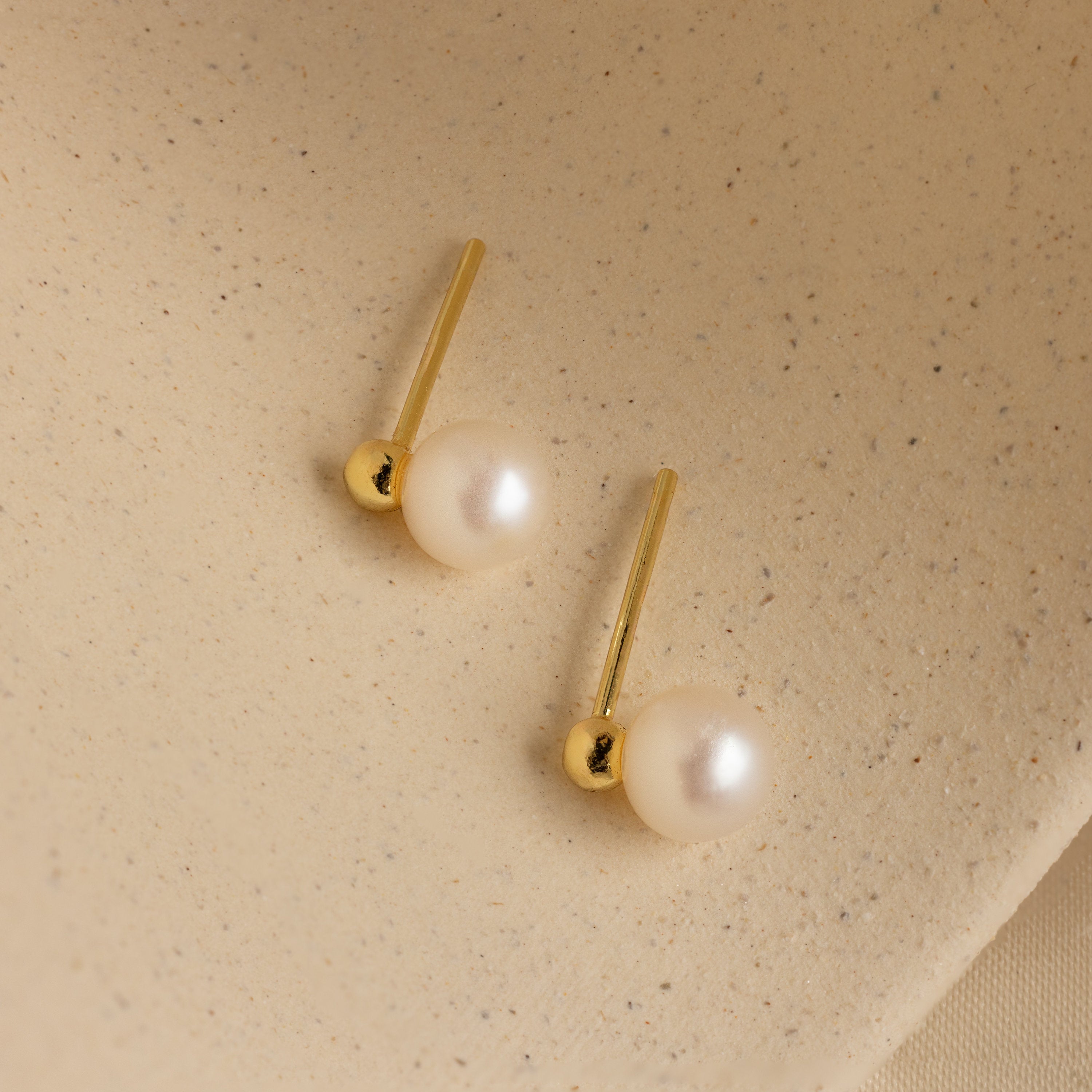 Classic Pearl Studs displayed on a textured beige surface, perfect for those who appreciate dainty earrings with timeless elegance.