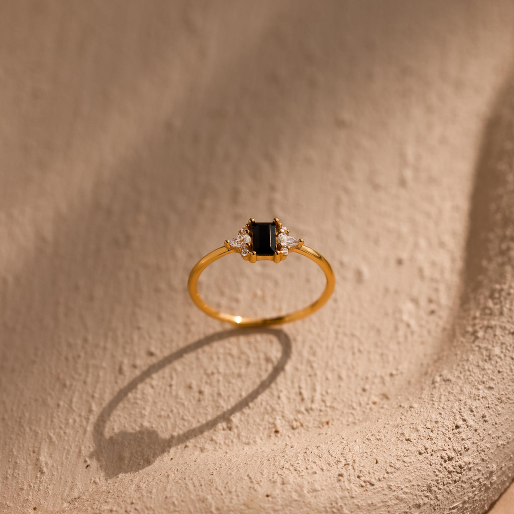 The Black Baguette Diamond Ring in 18K Gold features a rectangular black stone and small clear stones, showcased on a textured beige surface—a stunning statement piece with vintage style appeal.