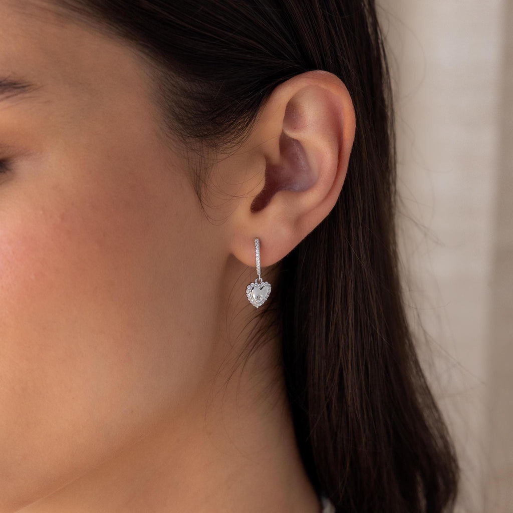 Close-up of a woman's ear wearing Pave Heart Drop Huggies—silver, heart-shaped huggie earrings adorned with sparkling gemstones, perfect as a romantic gift.
