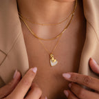 A woman wears layered gold necklaces, featuring the Stylish Pearl Initial Heart Necklace, over a beige blazer.
