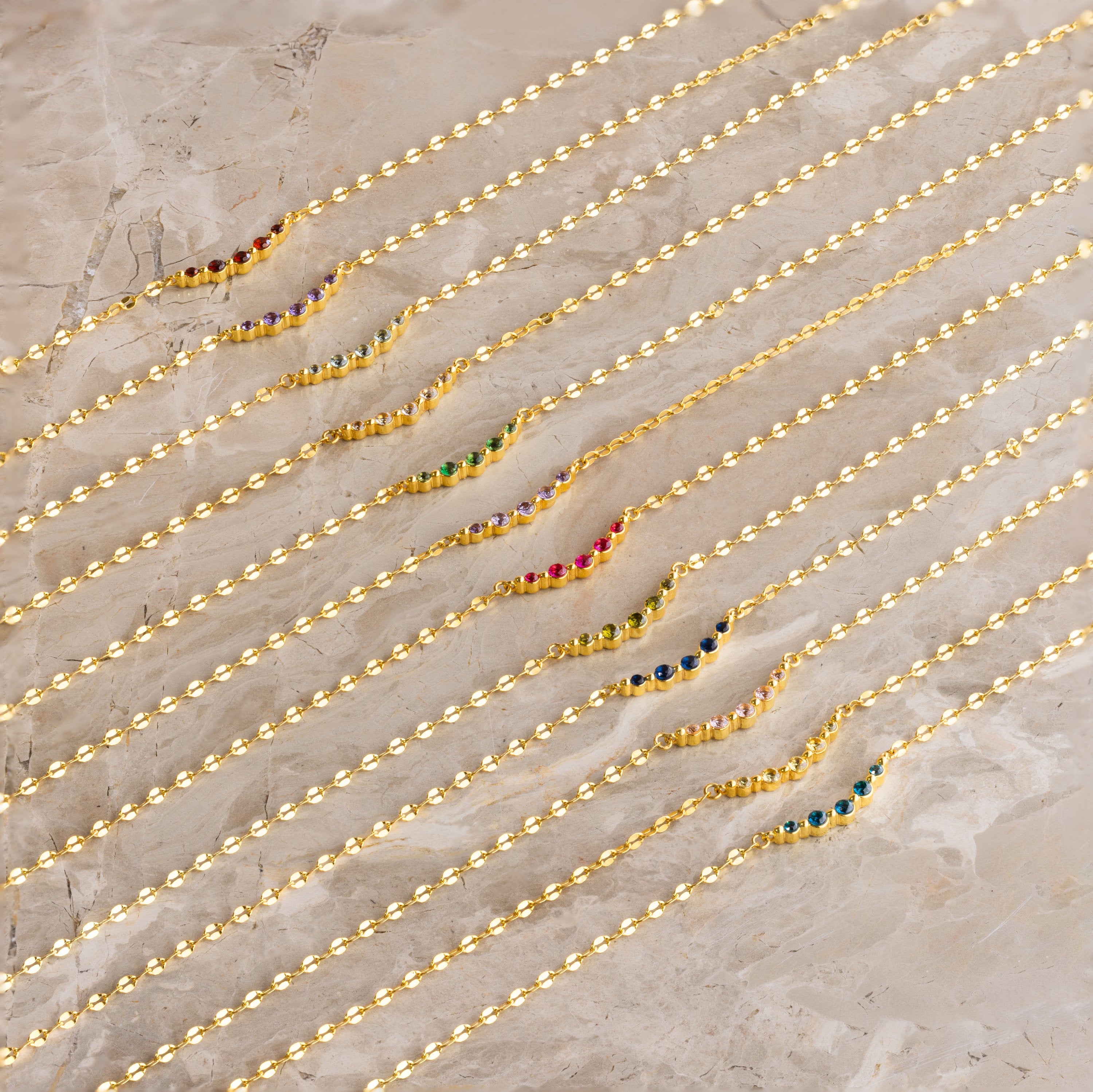 Nine Lace Birthstone Necklaces, each featuring a delicate gold chain and colorful gemstone, are arranged diagonally on a marble surface—perfect as dainty, personalized jewelry.