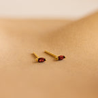 Two gold marquise-cut stud earrings with vibrant red gemstones from our Marquise Birthstone Studs collection, perfect for adding a personalized touch to your look. Displayed on a sandy beige surface.