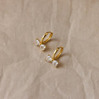 Diamond Bow Huggies, gold hoop earrings inspired by delicate bowties and adorned with sparkling gemstones, displayed on a crumpled beige background.