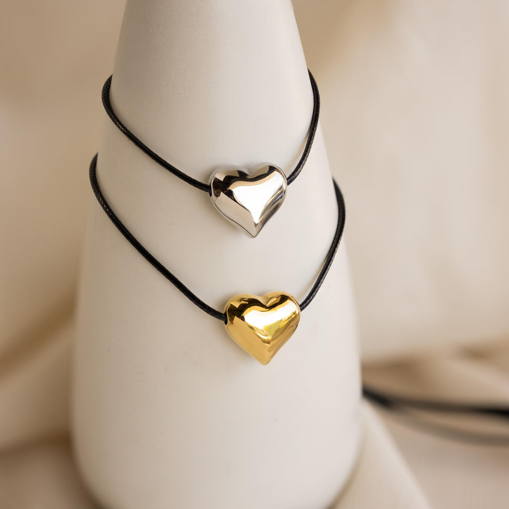 Two black cord bracelets with metallic heart charms—one silver, one gold—displayed on a white stand. Pair them with the Chunky Puff Heart Necklace for a coordinated look.