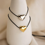 Two black cord bracelets with metallic heart charms—one silver, one gold—displayed on a white stand. Pair them with the Chunky Puff Heart Necklace for a coordinated look.