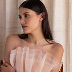 A woman with dark hair wears an off-shoulder, ruffled pale dress and the Herringbone Bow Necklace, capturing a soft coquette look as she gazes to the side.