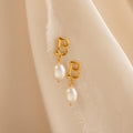 The Initial Pearl Drop Studs feature gold studs with letter B charms and dangling white pearls, displayed on a soft beige fabric background.