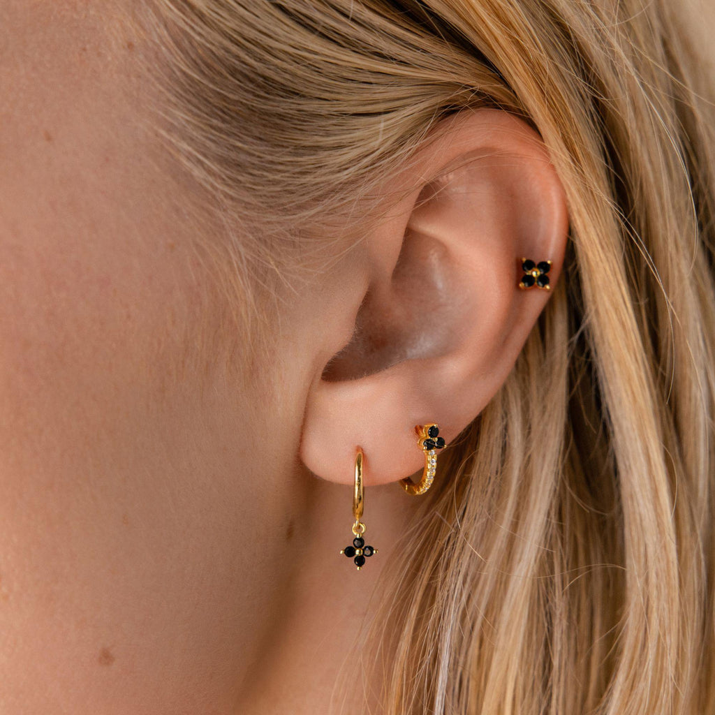 Close-up of a woman’s ear wearing three gold earrings, including the Black Ivy Flower Huggies with unique floral design elements and small black stones.