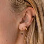 Close-up of a woman’s ear wearing three gold earrings, including the Black Ivy Flower Huggies with unique floral design elements and small black stones.