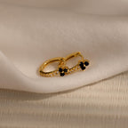The Onyx Lily Huggies are gold hoop earrings adorned with small black stones and diamonds, shown on soft white fabric for an elegant, dainty appearance.
