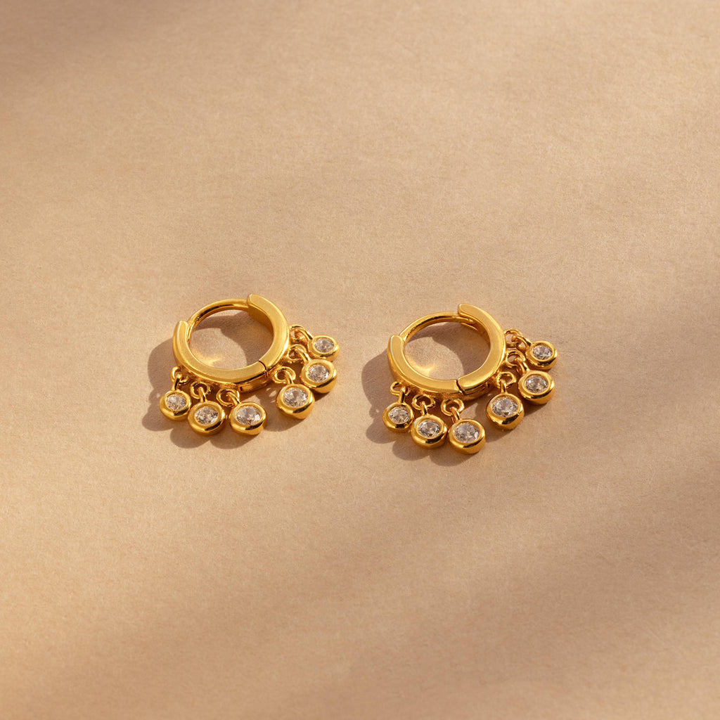 Chandelier Diamond Huggies—gold hoop earrings with dangling clear stones inspired by art deco elegance—displayed on a beige background.