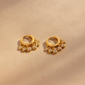 Chandelier Diamond Huggies—gold hoop earrings with dangling clear stones inspired by art deco elegance—displayed on a beige background.