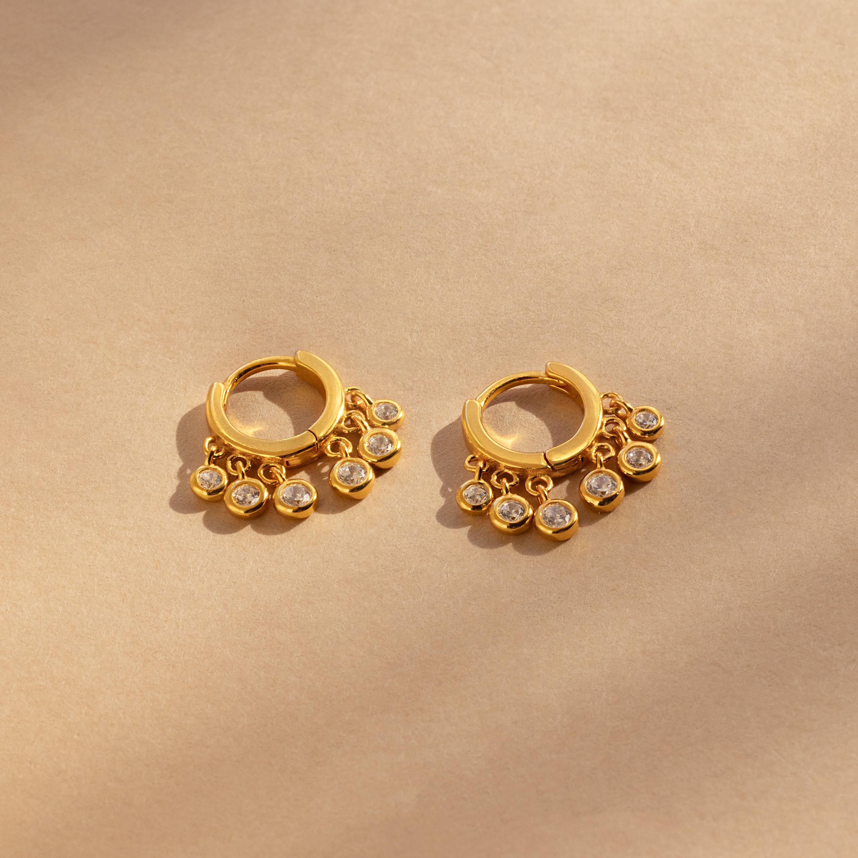 Chandelier Diamond Huggies—gold hoop earrings with dangling clear stones inspired by art deco elegance—displayed on a beige background.