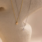 The Tiny Rose Quartz Heart Necklace, shown alongside gold and silver heart-shaped pendant necklaces, is displayed on a textured beige surface.