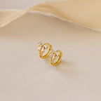Close-up of Diamond Ribbon Bow Huggies in Gold set against a cream background.