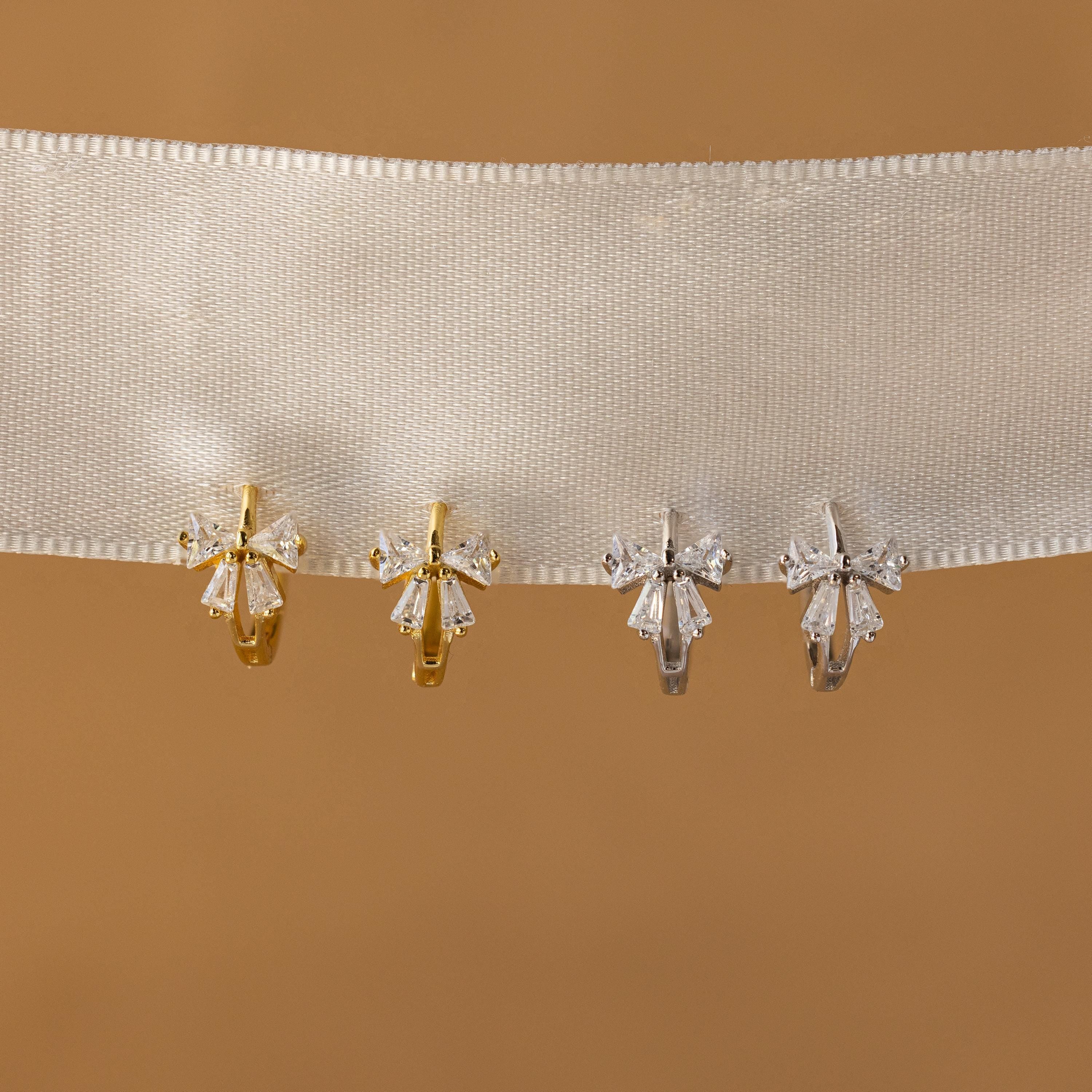 Four pairs of Diamond Ribbon Bow Huggies in 18K Gold, two gold and two silver, displayed on a white ribbon against a tan background.