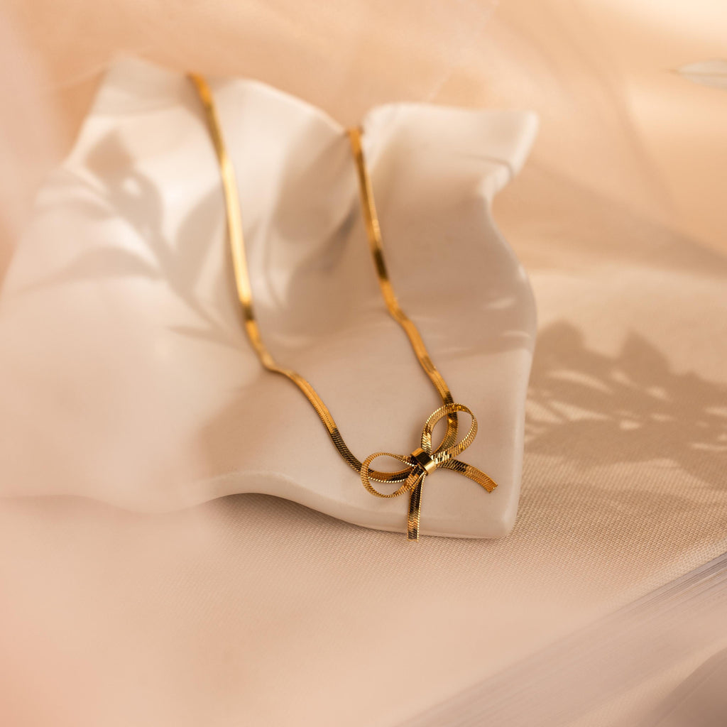 Herringbone Bow Necklace in coquette style, displayed on a wavy white dish with soft, warm lighting and shadows.