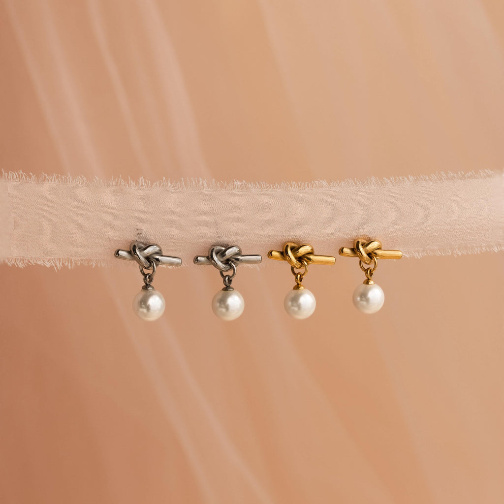 Two pairs of Love Knot Pearl Earrings—one silver, one gold—displayed on sheer beige fabric. Ideal as a wedding accessory or for anyone who loves vintage-inspired heart earrings.