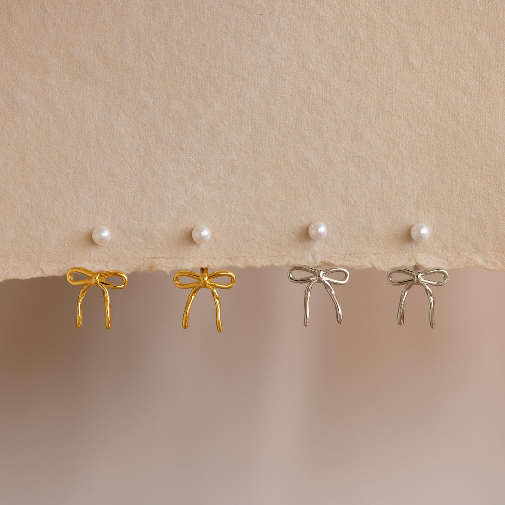 Two pairs of Pearl & Bow Ear Jackets in 18K Gold—one in gold and one in silver—are displayed on a textured beige surface with pearl stud earrings above.
