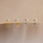 Two pairs of Pearl & Bow Ear Jackets in 18K Gold—one in gold and one in silver—are displayed on a textured beige surface with pearl stud earrings above.