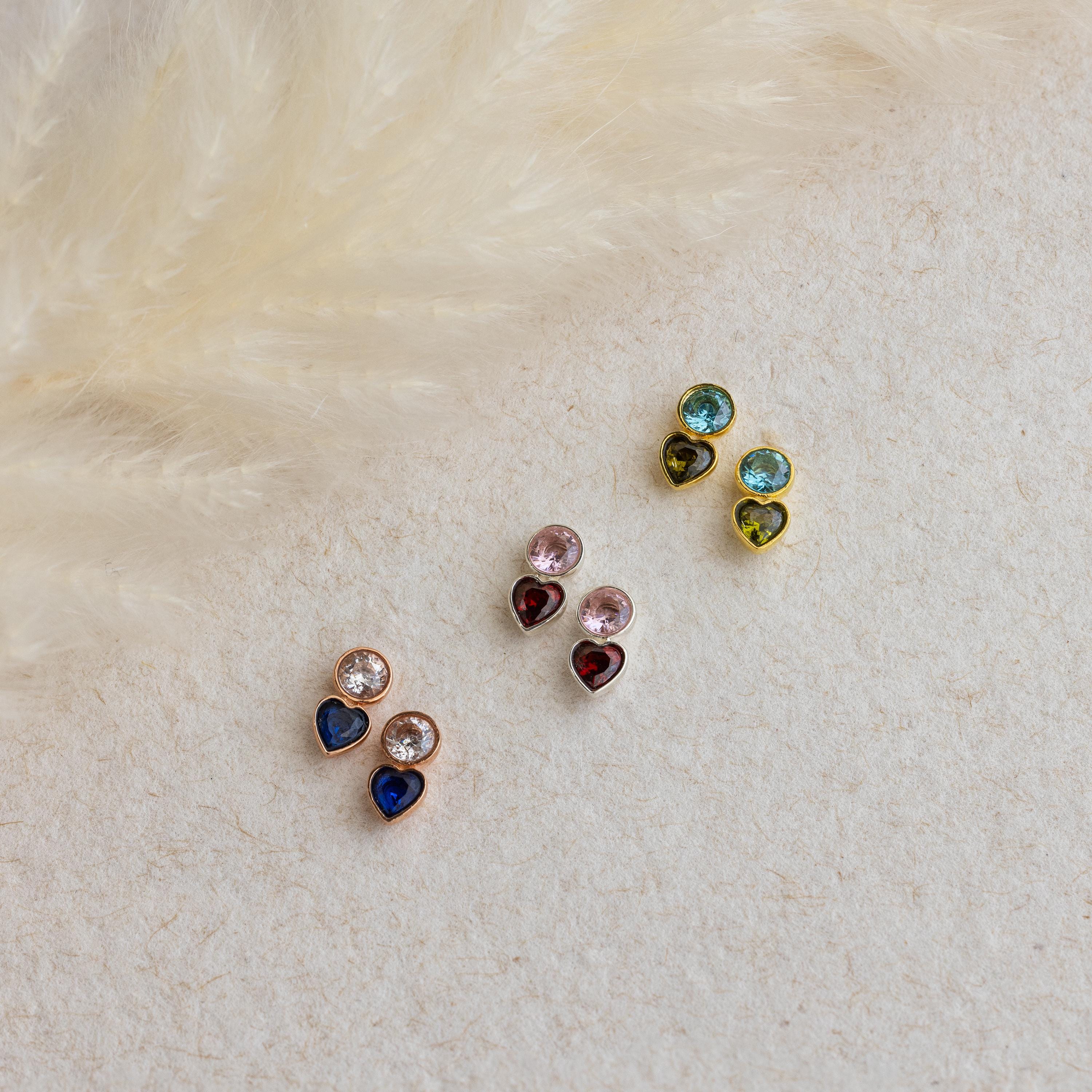 The Toi et Moi Birthstone Studs, along with three other heart-shaped and round gemstone earring pairs, are displayed on a textured surface near soft beige feathers—ideal as a personalized gift or custom stud earrings.