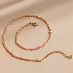 The Blush Pearl Beaded Necklace features pink freshwater pearls with a gold clasp and extender, elegantly displayed on a beige background.