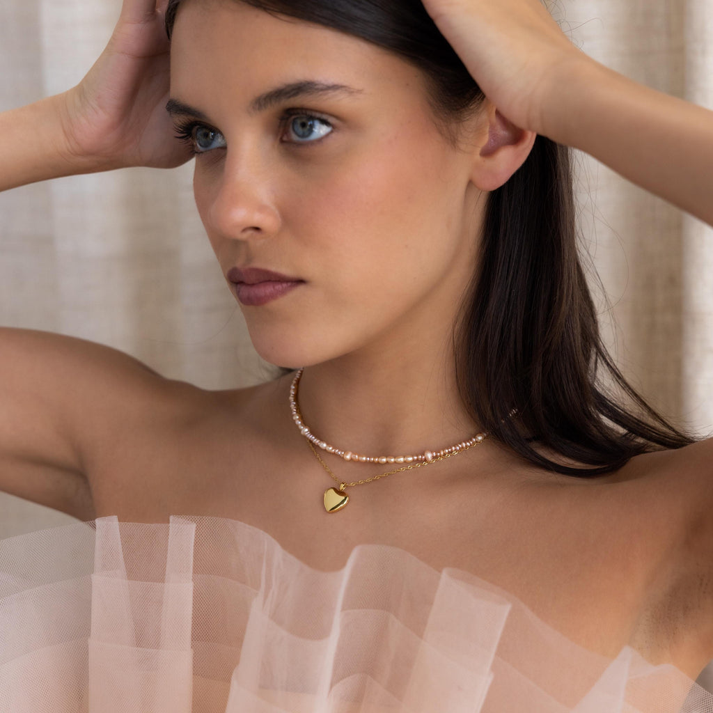 A woman in a sheer, ruffled top wears the Blush Pearl Beaded Necklace featuring a gold heart pendant, with her hands gently touching her head.