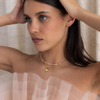 A woman in a sheer, ruffled top wears the Blush Pearl Beaded Necklace featuring a gold heart pendant, with her hands gently touching her head.