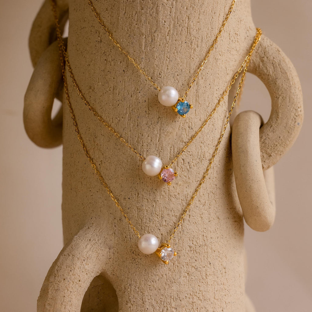 The Pearl & Birthstone Necklace, featuring pearls and colorful gemstones, is elegantly displayed draped over a textured beige sculpture.