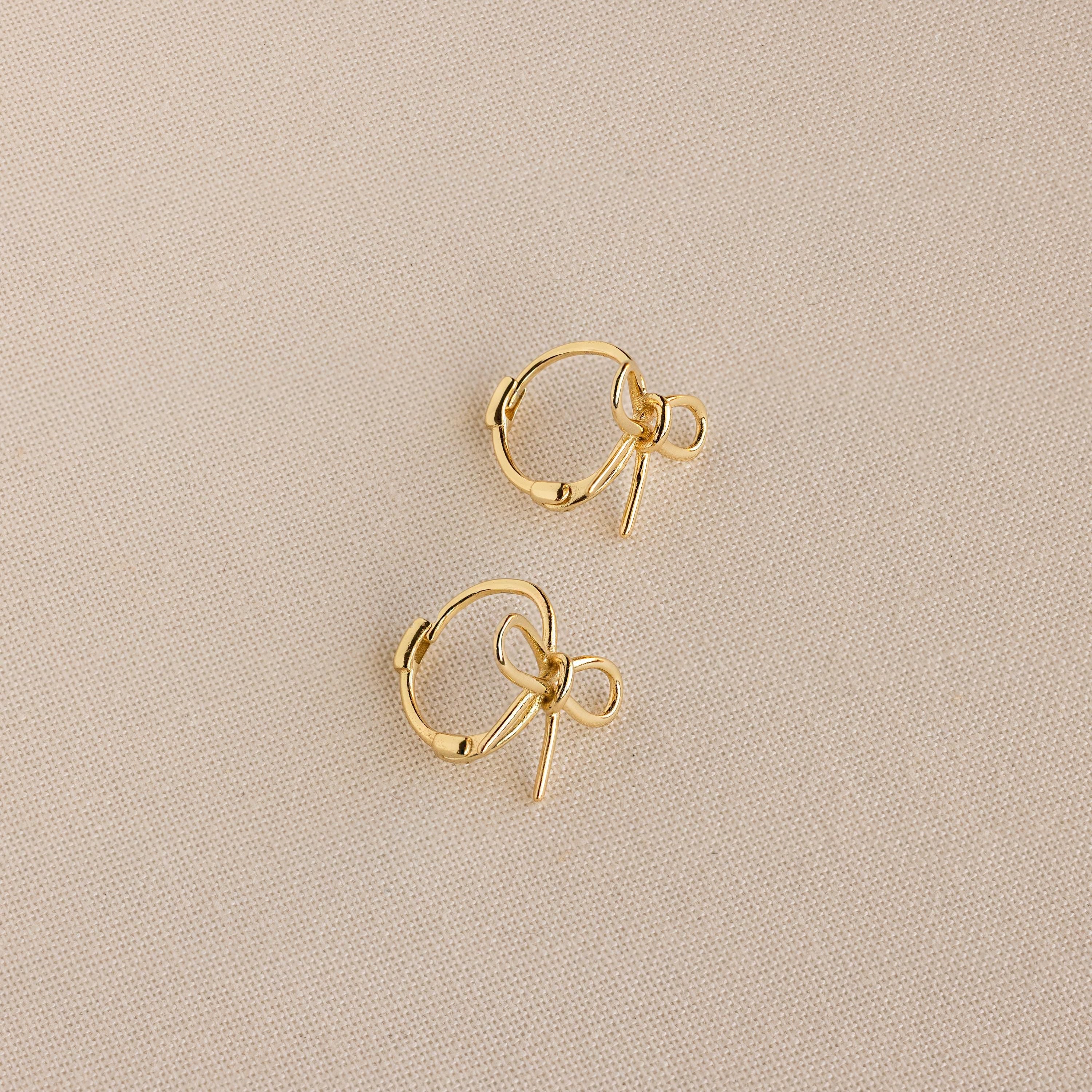 The Classic Ribbon Bow Huggies are small gold hoops with delicate ribbon bow charms, shown on a beige textured fabric background, adding whimsical femininity to any outfit.