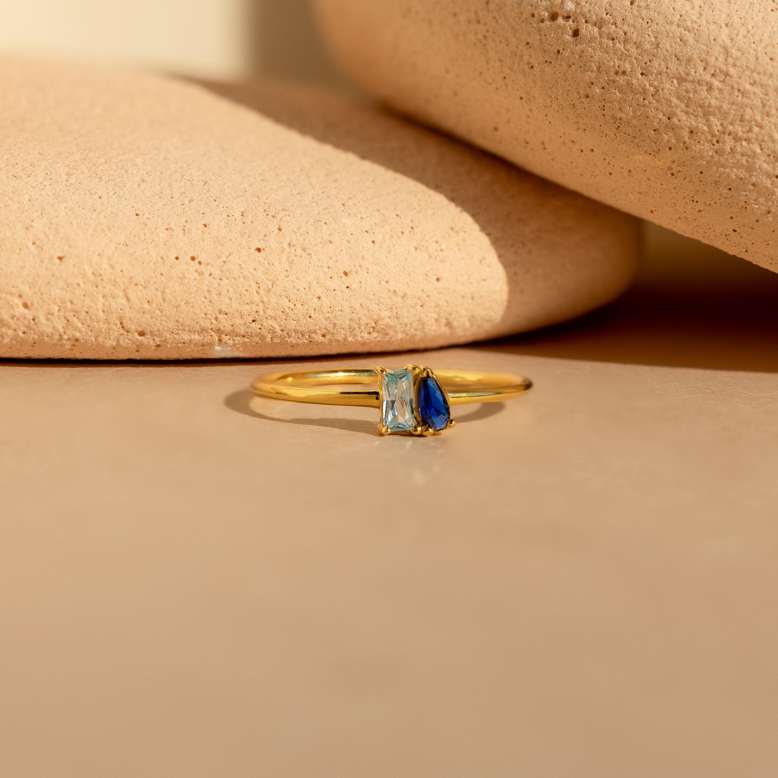 The Tiny Toi et Moi Birthstone Ring features a rectangular clear stone and a teardrop-shaped blue stone, beautifully displayed on a beige surface with stone accents.