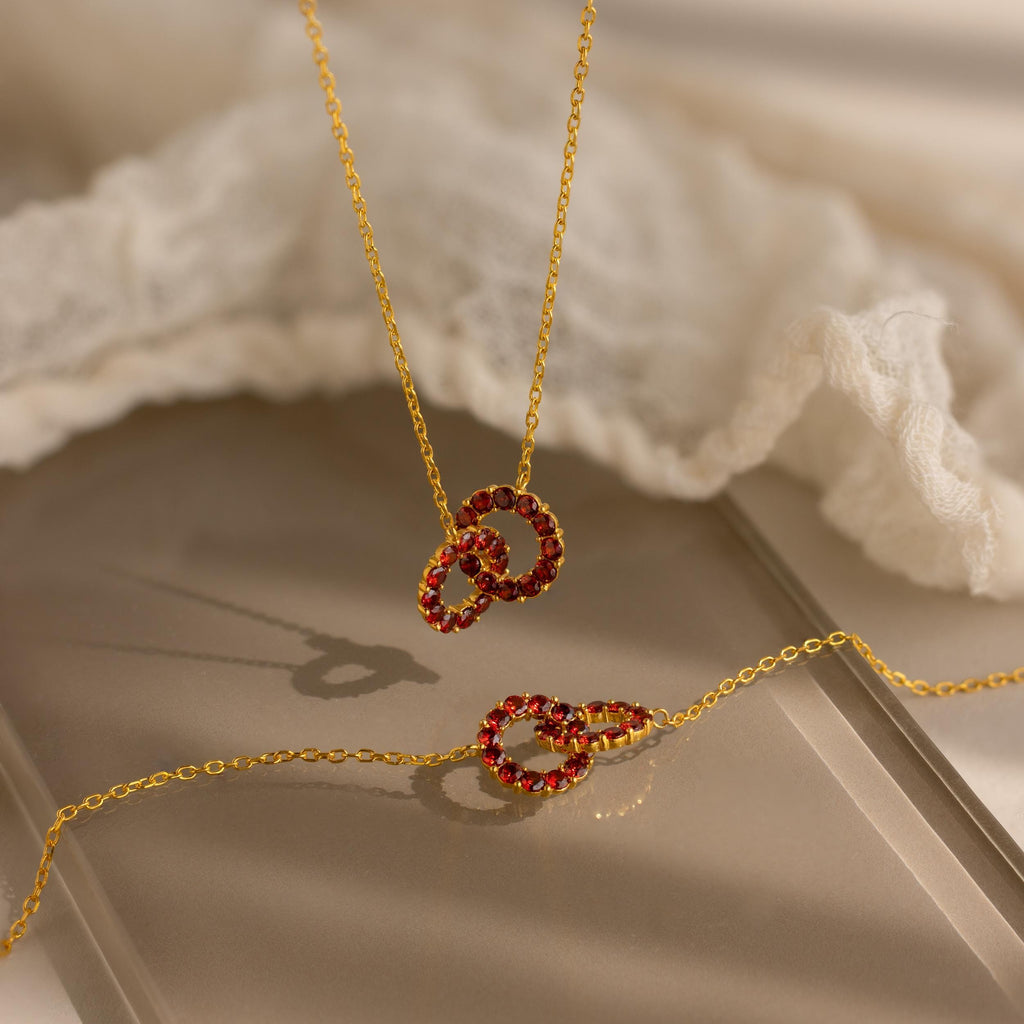 Gold necklaces with red gemstone interlocking circle pendants displayed on neutral fabric, inspired by the Interlocking Duo Birthstone Bracelet—a meaningful, customizable birthstone gift for someone special.