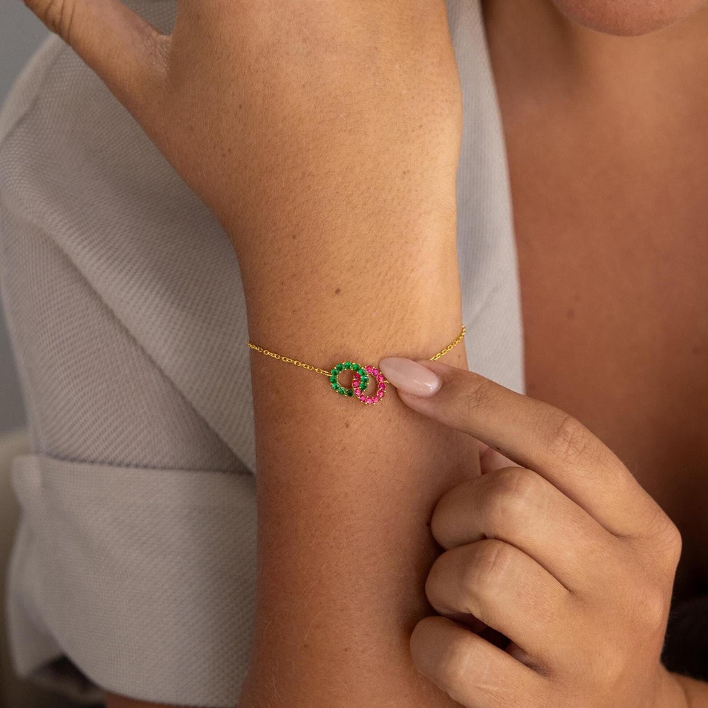 A person points to the Interlocking Duo Birthstone Bracelet on their wrist, highlighting its customizable design with two interlocking rings—a green and a pink—making it a meaningful gift.