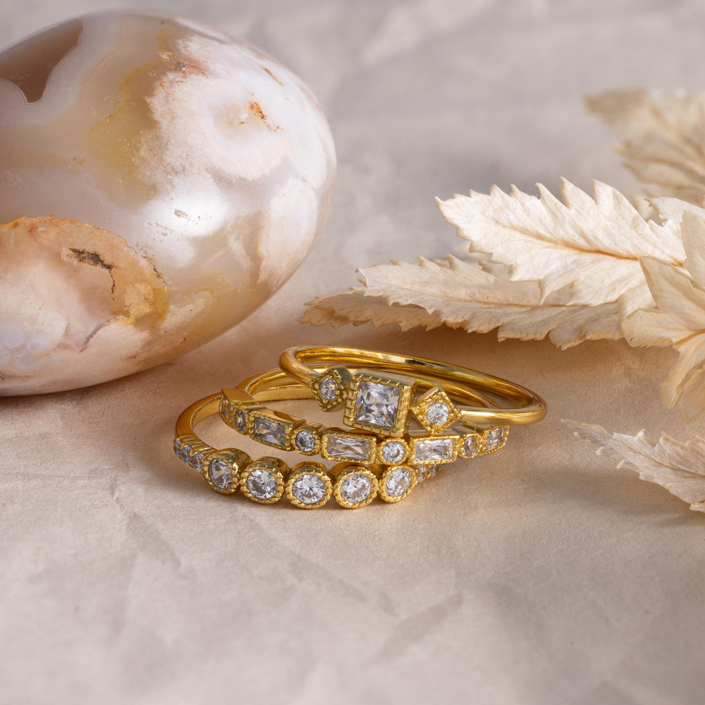 Stacked gold Art Deco diamond rings displayed on a textured surface beside a polished agate stone and dried leaves.