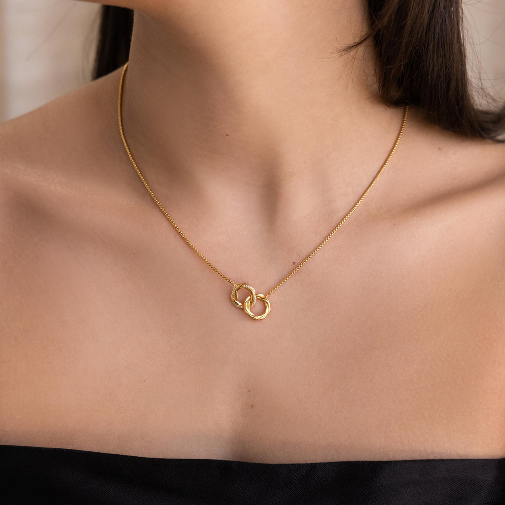 A woman in a strapless top is shown from the shoulders up wearing the Interlocking Pave Charm Necklace, which features two interlocking diamond-encrusted rings.