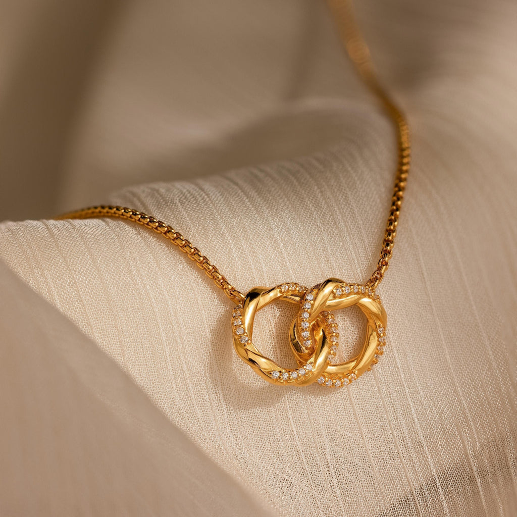 Interlocking Pave Charm Necklace featuring two interlocked rings—one adorned with diamonds—elegantly displayed on a cream fabric background.