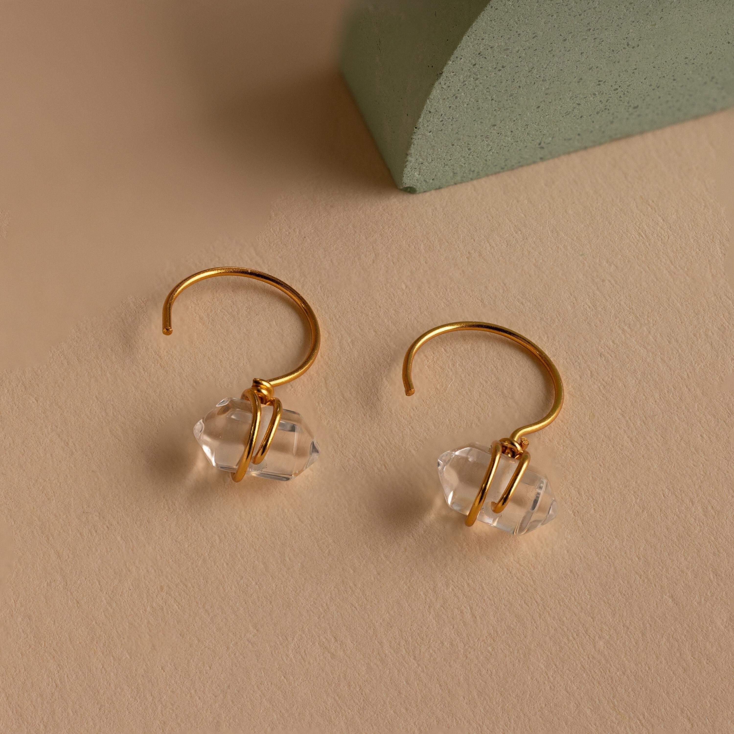 The Herkimer Diamond Threaders, gold hoop earrings with clear crystal pendants inspired by nature, are displayed on a beige surface beside a green block.