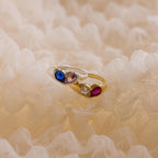 The Duo Birthstone Heart Signet Ring, crafted in gold and adorned with blue, clear, and red gemstones, rests on soft cream-colored fabric.